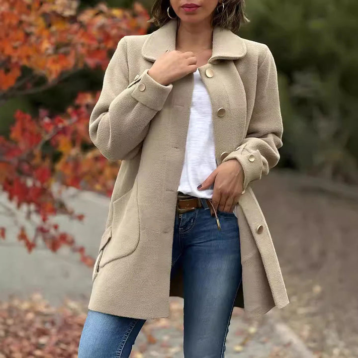 Woman wearing a beige coat and blue jeans with a blurred autumn background